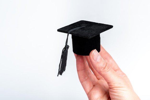 Una mano che tiene un berretto in miniatura di un laureato di un college universitario di scuola superiore su uno sfondo bianco spazio di copia Cappello di laurea nero con nappa nera