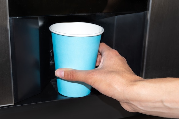 Una mano che tiene un bicchiere di carta blu per versare una bevanda da un distributore automatico. Versare acqua, caffè in un bicchiere di carta