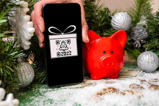 Una mano che tiene uno smartphone con un simbolo regalo e un codice QR a forma di abete, un salvadanaio rosso su monete d'oro coperte di neve e rami di abete natalizio. Shopping online di Natale