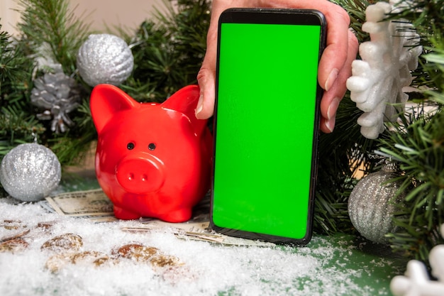 Una mano che tiene uno smartphone con uno schermo verde vuoto, un salvadanaio rosso su monete d'oro e banconote coperte di neve e rami di abete verde natalizio decorati con palline.