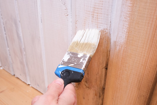 Una mano dipinge una parete di superficie in legno con vernice acrilica bianca, riparazione e restauro della casa.