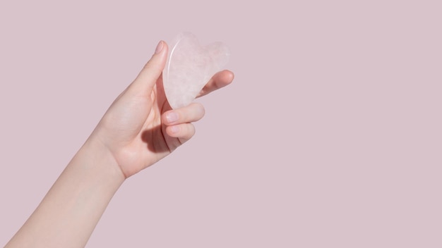 Una mano femminile che tiene il massaggiatore viso gua sha su sfondo rosa