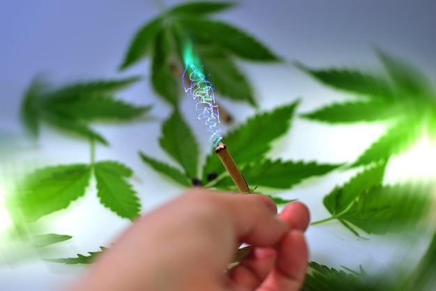 Una mano femminile con una sigaretta accesa con fuoco elettrico astratto su uno sfondo di foglie di cannabis