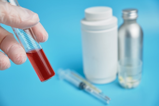 Una mano in guanti detiene una provetta con test rossi sullo sfondo di lattine mediche, pillole su sfondo blu.