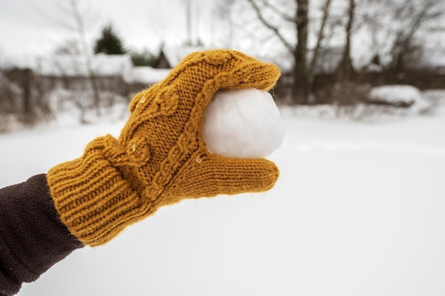 Una mano in un guanto giallo tiene una palla di neve nel cortile contro un orto innevato