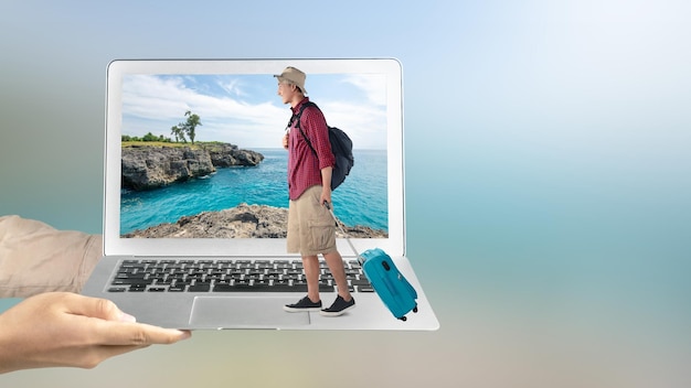 Una mano umana che tiene un portatile con un uomo asiatico con un cappello e uno zaino che porta una valigia in piedi con una scogliera marina e una vista sull'oceano sullo schermo del portatile
