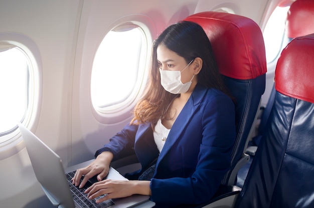 Una maschera facciale da portare della giovane donna di affari sta usando il laptop a bordo, nuovo viaggio normale dopo il concetto di pandemia covid-19