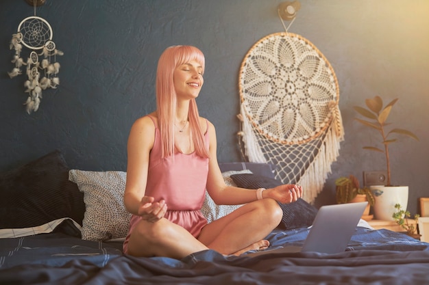 Una modella femminile abbastanza sorridente medita vicino al laptop su un letto grande vicino a un muro blu scuro