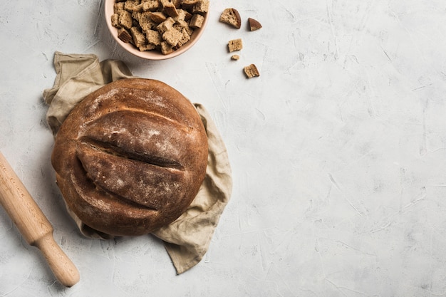 Una pagnotta rotonda si trova su un tovagliolo di lino grigio con pangrattato in una ciotola e un mattarello per impasto su uno sfondo chiaro.