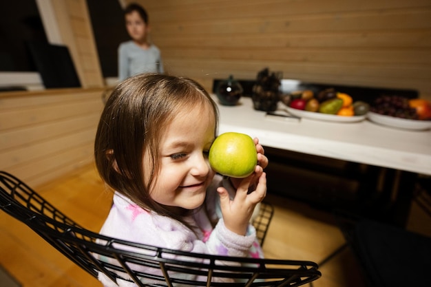 Una piccola bambina carina in pigiama si siede in cucina con mela verde in un'accogliente casetta in legno La vita in campagna