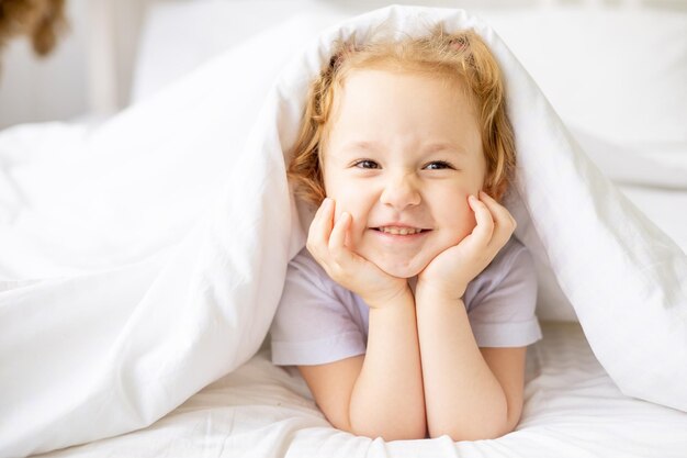 Una piccola bambina carina su un letto di cotone bianco a casa si concede e si diverte a sorridere sul letto di casa con le mani piegate sotto le guance
