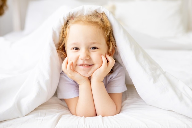 Una piccola bambina carina su un letto di cotone bianco a casa si concede e si diverte a sorridere sul letto di casa con le mani piegate sotto le guance