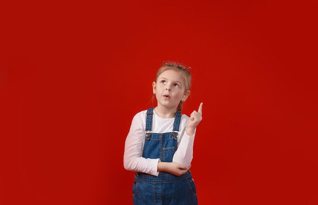 Una piccola bella studentessa in un vestito di jeans posa su uno sfondo rosso isolato