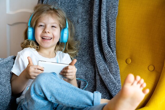 Una piccola bionda felice a casa su una sedia che ascolta musica con le cuffie e suona