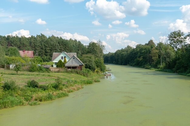 Una piccola casa in legno in un villaggio sulla riva del fiume Foresta in fiore lungo la riva del fiume e nuvole sull'acqua in una soleggiata giornata estiva