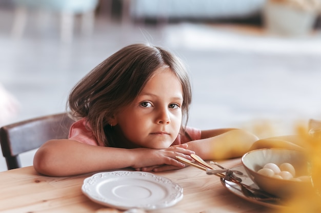 Una piccola dolce ragazza siede pensierosa al tavolo della cucina. Bambino sognante