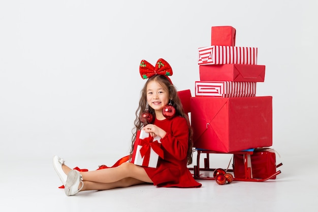 Una piccola ragazza asiatica seduta in un vestito rosso si siede con scatole regalo su uno sfondo bianco. Concetto di Natale, spazio di testo