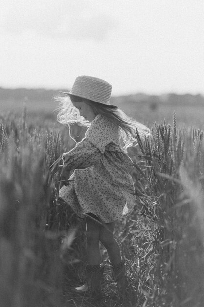 Una piccola ragazza attraente con un cappello di paglia corre su un campo di grano con le orecchie verdi