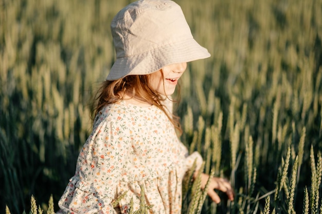 Una piccola ragazza attraente con un cappello di paglia corre su un campo di grano con le orecchie verdi