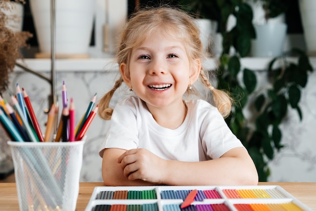 Una piccola ragazza bionda con un sorriso affascinante si siede a un tavolo con una tavolozza di colori vivaci