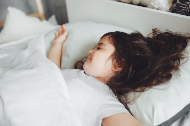 Una piccola ragazza bruna sta dormendo nel suo letto con lo spazio della biancheria da letto bianca per un sonno sano del bambino di testo