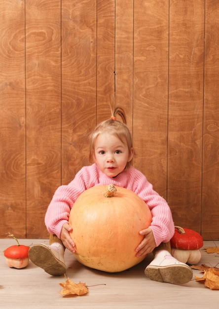 Una piccola ragazza carina con un maglione rosa abbraccia una grande zucca seduta sul pavimento su un legno.
