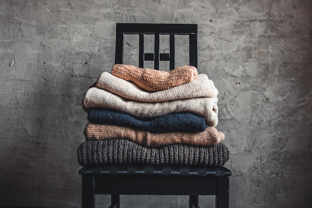Una pila di maglioni caldi e accoglienti lavorati a maglia, su una sedia vicino al muro grigio. autunno, concetto di inverno.