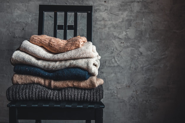 Una pila di maglioni caldi e accoglienti lavorati a maglia, su una sedia vicino al muro grigio. autunno, concetto di inverno.