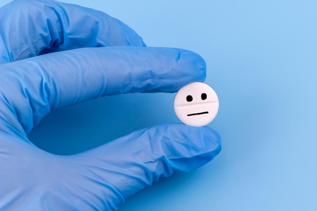 Una pillola con un'emoticon triste in una mano in un guanto medico su sfondo blu Assistenza sanitaria