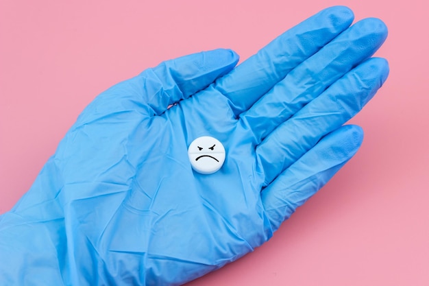 Una pillola con un'emoticon triste in una mano in un guanto medico su sfondo rosa Assistenza sanitaria