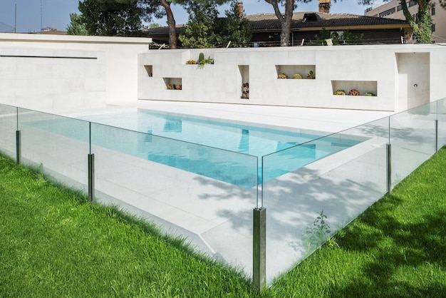 Una piscina in marmo bianco con una ringhiera in vetro, un giardino con prato verde e nicchie con vasi di fiori in una parete
