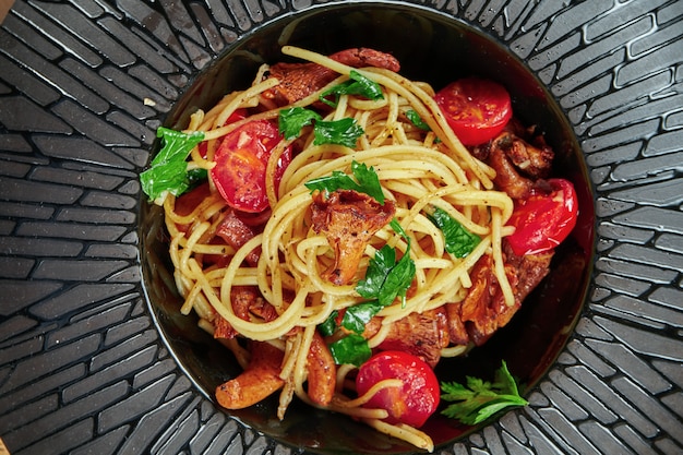 Una porzione appetitosa di pasta italiana fatta in casa (spaghetti) con funghi di bosco (finferli) pomodorini e rucola in un piatto nero su un tavolo di legno. Tavolo da ristorante