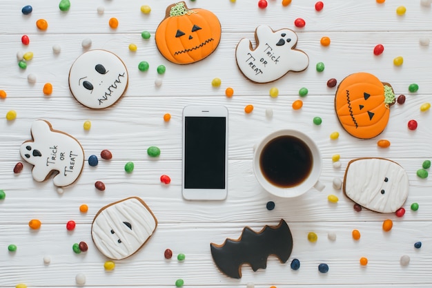 Una preparazione per Halloween: tazza di caffè e smartphone sul fondo bianco in legno con caramelle e panpepato.