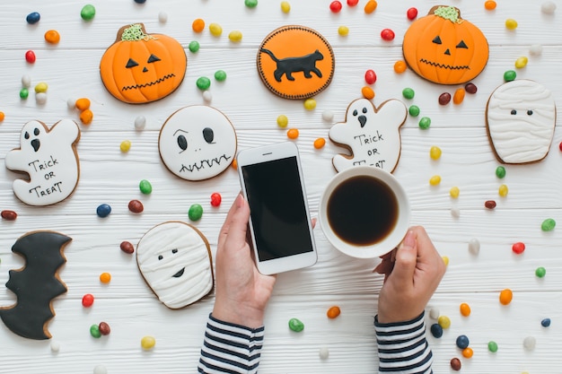 Una preparazione per Halloween. Una ragazza tazza di caffè e smartphone sul fondo bianco in legno con caramelle e panpepato.