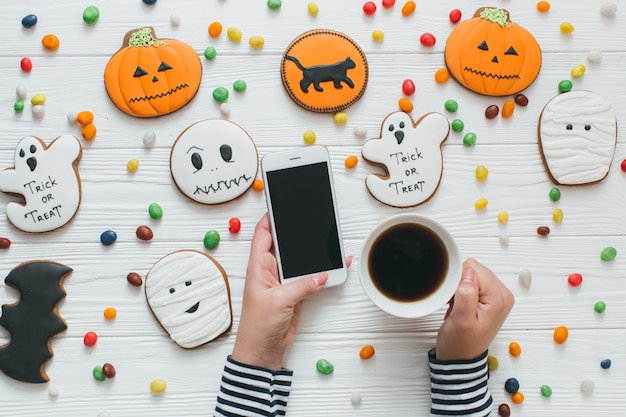 Una preparazione per Halloween. Una ragazza tazza di caffè e smartphone sul fondo bianco in legno con caramelle e panpepato.