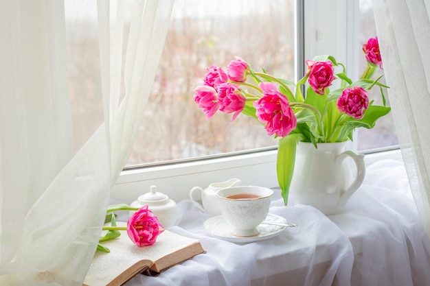 Una primavera ancora in vita un mazzo di tulipani in un vaso una tazza di tè e un vecchio libro giacciono sulla finestra