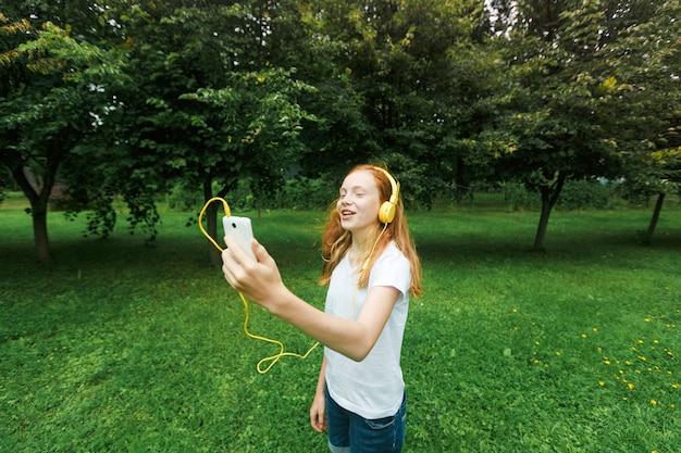 Una ragazza adolescente con i capelli rossi ascolta musica con le cuffie in un parco in estate. Una giovane donna gode e si rilassa all'aperto in una giornata di sole.