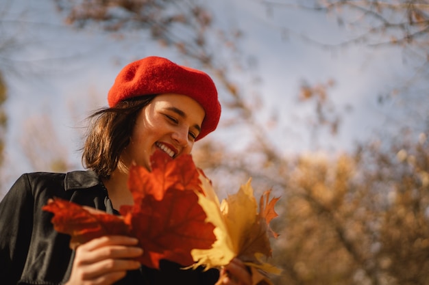 Una ragazza adolescente con un berretto rosso con un mazzo di foglie autunnali in mano cammina attraverso la foresta