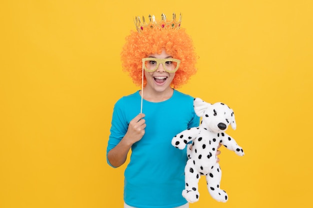 Una ragazza adolescente felice con una parrucca da clown fantasia indossa la corona della regina e gli occhiali da festa divertenti tengono l'infanzia del giocattolo