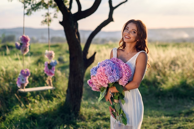 Una ragazza affascinante con un bel sorriso in un abito bianco con un mazzo di teneri fiori colorati al tramonto.