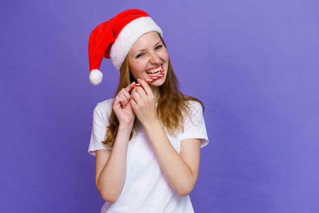 Una ragazza allegra con un cappello da Babbo Natale sta mangiando lecca-lecca e ride in maglietta bianca caucasica giovane donna e...