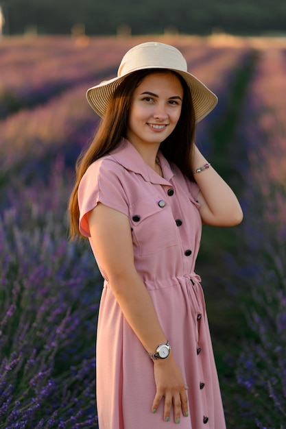 Una ragazza allegra con un vestito rosa e un cappello di paglia tra le mani si trova tra i cespugli di lavanda