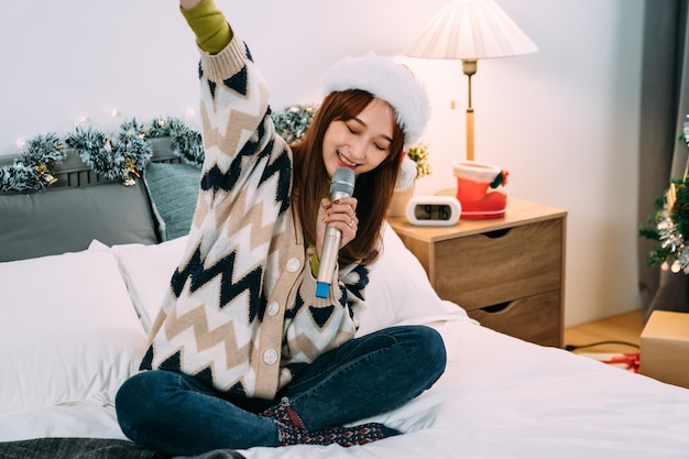 una ragazza asiatica allegra che celebra il Natale da sola sta alzando il braccio mentre si diverte a cantare il karaoke sul letto nella camera da letto di casa.