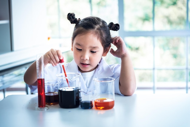 Una ragazza asiatica di 4 anni che indossa un'uniforme da scienziato bianco Apprendimento e condusse un esperimento scientifico su un tavolo bianco con un misurino e una provetta posizionati, ai bambini e al concetto di istruzione.