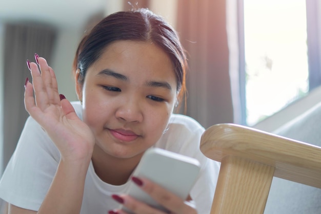 Una ragazza asiatica felice dice "ciao" alla telecamera su una videochiamata sullo smartphone mentre si rilassa sul divano a casa