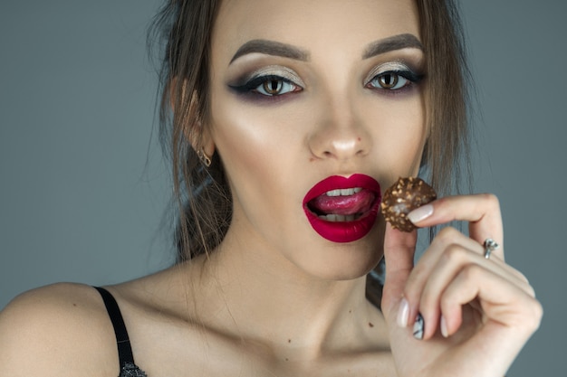 Una ragazza attraente con le labbra rosse mangia caramelle al cioccolato e guarda la telecamera in studio