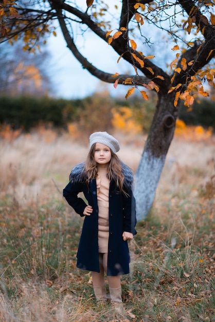 Una ragazza attraente con un cappotto blu scuro e un cappuccio grigio e un vestito beige in piedi nel giardino d'autunno