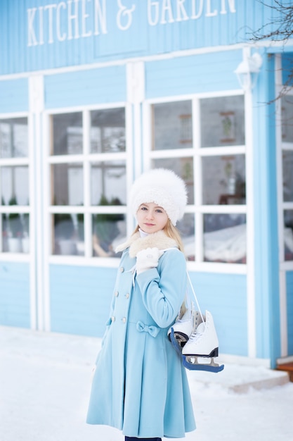 Una ragazza bionda carina andrà a pattinare all'aperto. Una ragazza in un cappotto blu e cappello di pelliccia con pattini a casa d'inverno. Attività del fine settimana con tempo freddo. Natale, concetto di vacanze invernali. Sport invernali.