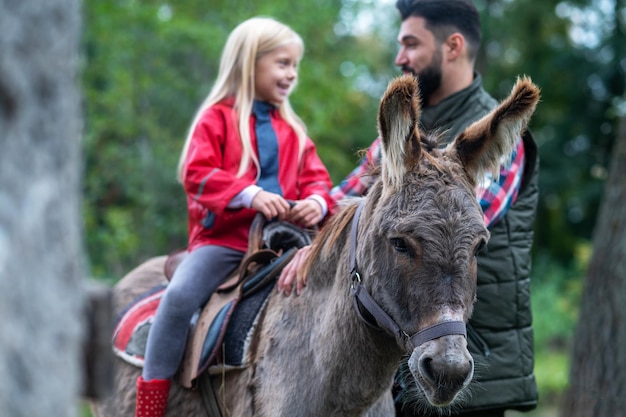 Una ragazza bionda carina cavalca un asino in una fattoria, suo padre l'aiuta