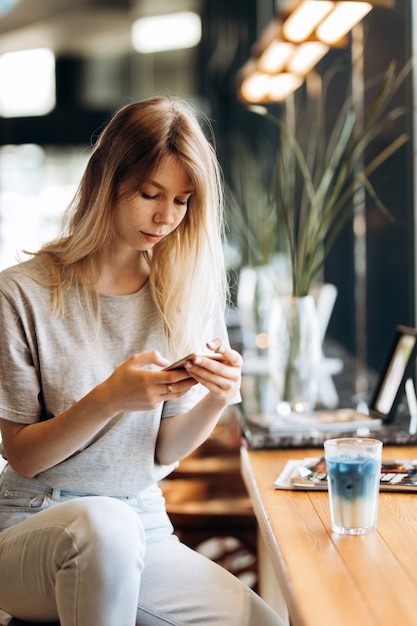 Una ragazza bionda carina e magra, vestita in stile casual, beve caffè e guarda il suo telefono in una caffetteria. .